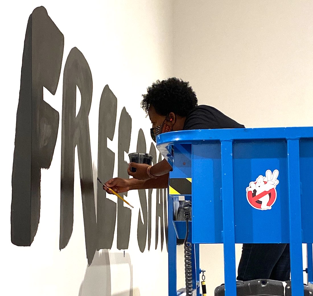 Artist Lisa McLymont is seen from the side, standing on a blue electric lift, paints the words "free space" in black on a white wall