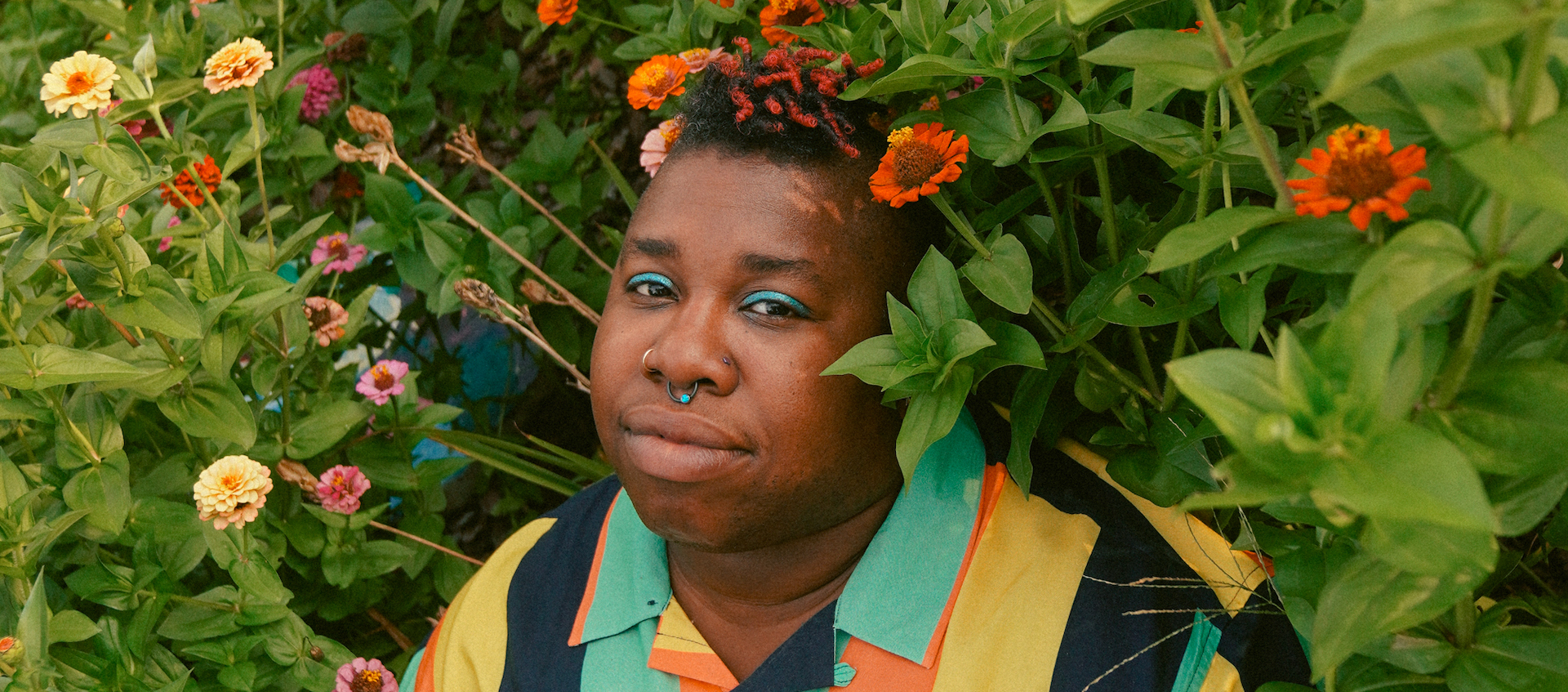 Singer-songwriter Sharon Udoh, wearing a bright collared shirt with vertical stripes, standing nestled in a large bush with orange and yellow flowers