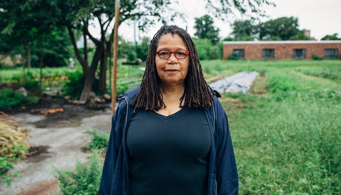 Artist and activist Linda Goode Bryant, wearing a dark blue shirt and glasses, stands outside in a grassy area with a vegetable garden to the side and a brick building in the distance