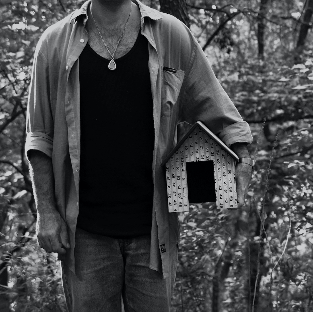 A black and white portrait of Cuban artist Juan-Si Gonzalez from the neck down, a birdhouse in the crook of his left arm