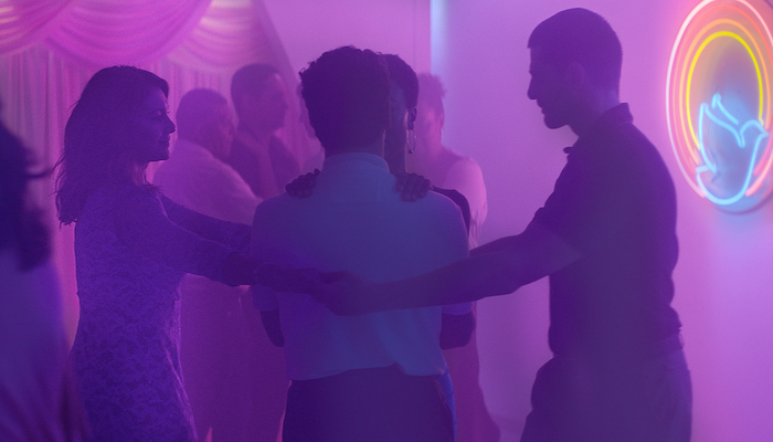 A husband and wife clasp their hands around another husband and wife couple in an embrace in a room lit by purple neon in a scene from Gabriel Mascaro's Divine Love