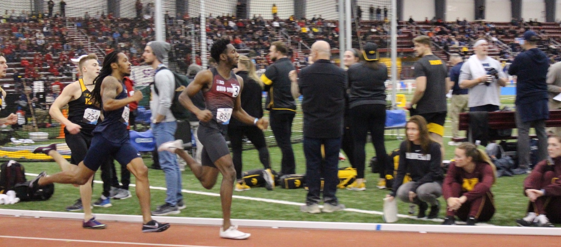 A group of college athletes run on an indoor track; a crowd is seen in the background