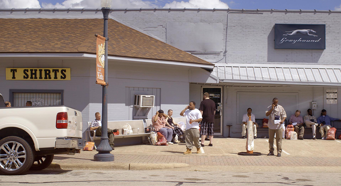 Image of a Greyhound station. People milling around in front of it, to the left a pick-up truck sits on the street