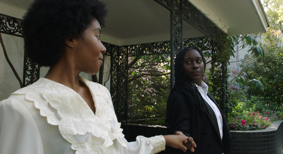 Two Black women stand next to one another, holding hands and looking into one another eyes. The woman on the left wears a white blouse and the woman on the right wears a black blazer