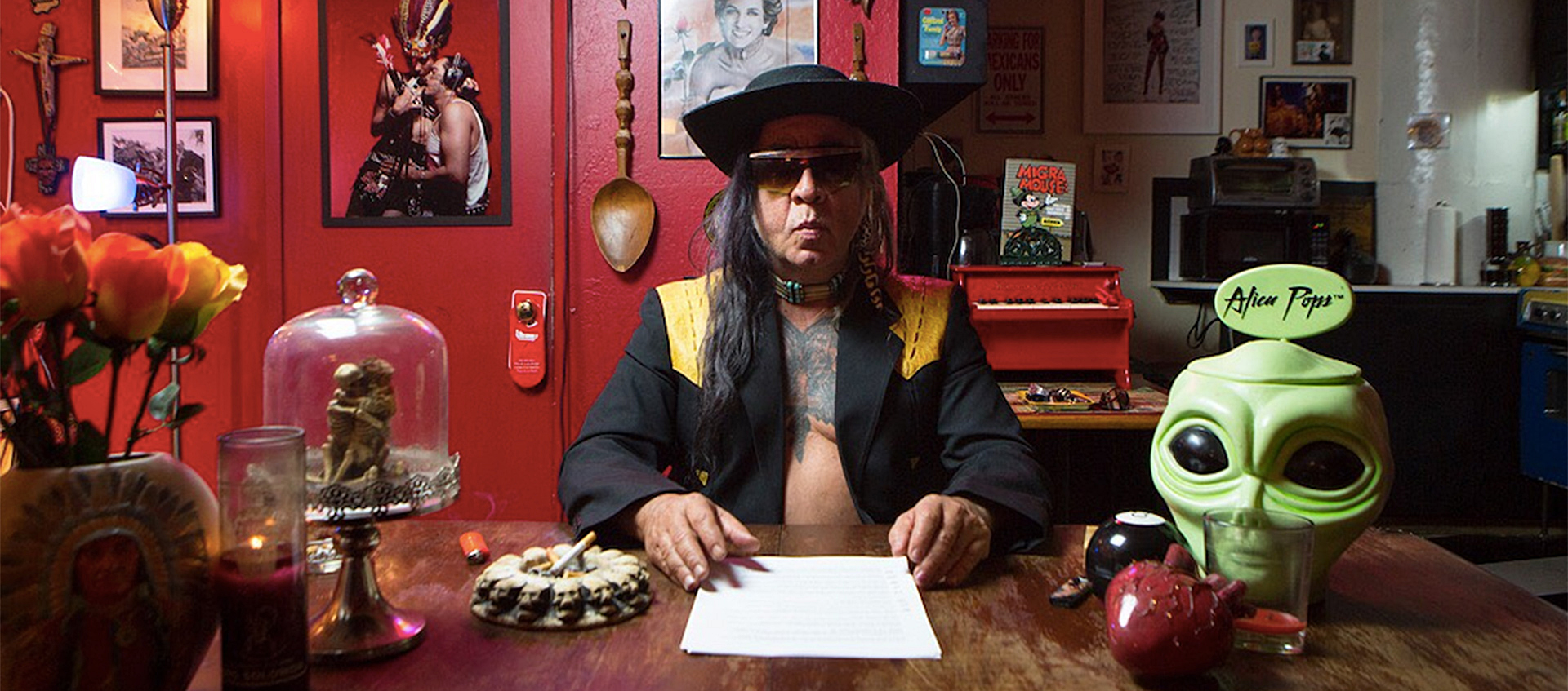 Gómez-Peña is seated in his home studio. He is wearing a black jacket with no shirt underneath. He wears a hat and in front of him is a piece of paper. To his right is a plastic, green alien head and to his left is an ashtray with a cigarette resting in it.