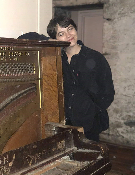 Image of Jo Snyder standing by an old piano. Jo is wearing a black shirt and rests their head on the top of the piano