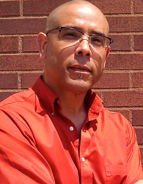 Maurice Stevens wearing glasses and an orange shirt standing in front of a brick wall