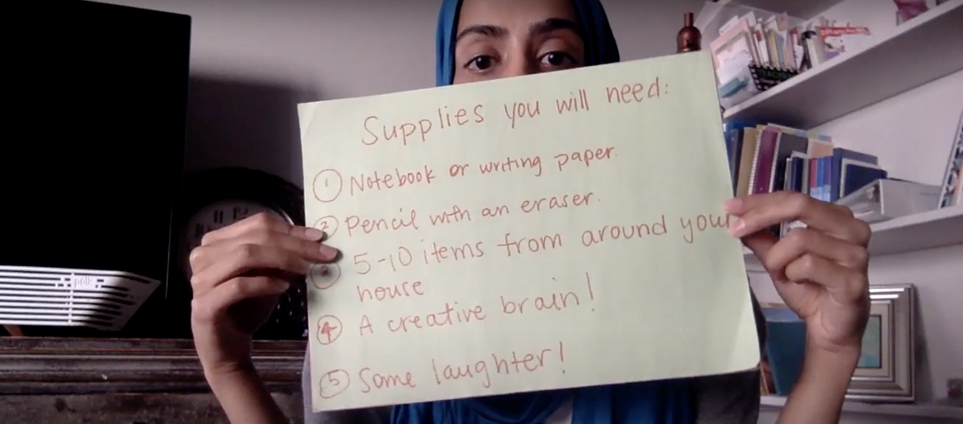 Artist Fariha Tayyab sits in her living room holding a small handwritten sign in front of her face, obscuring all of it but her eyes