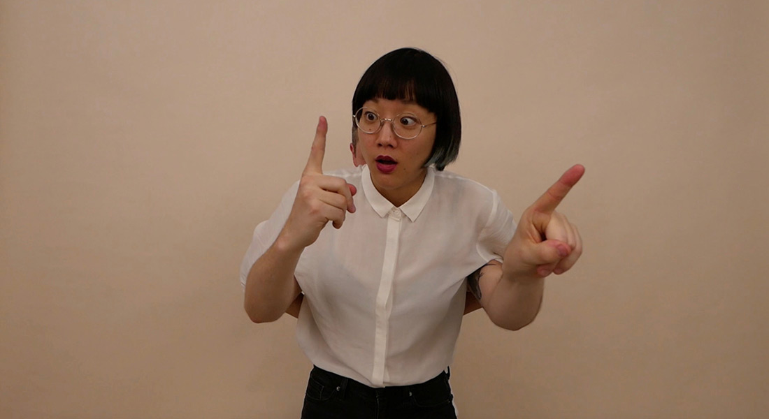 A person with black hair, eyeglasses, and a white shirt stands in front of a beige background. They are pointing the forefingers of their left and right hands to the ceiling