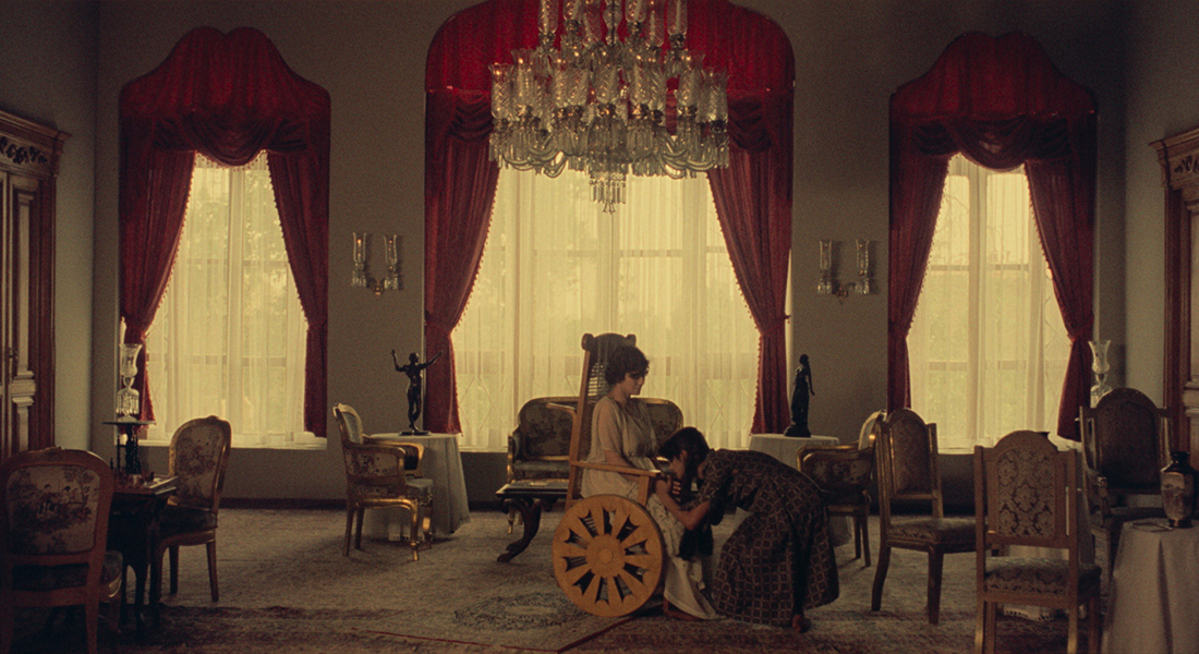 Two women in a large parlor room, one is seated in a wheelchair while another kneels in front of her