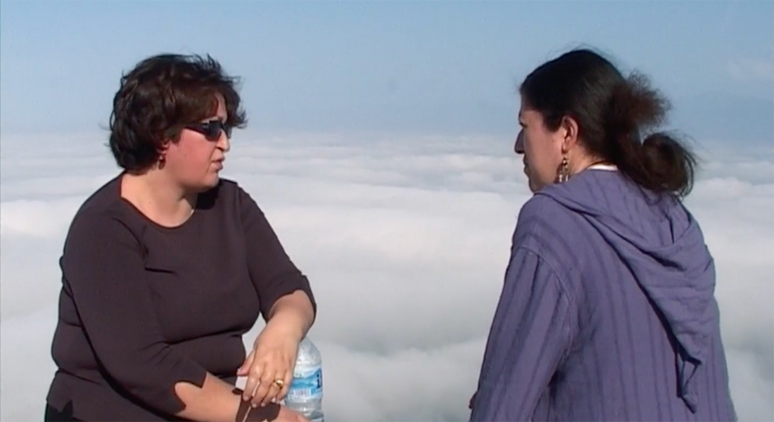 Two women having a conversation on a high altitude with clouds behind in the scene. 