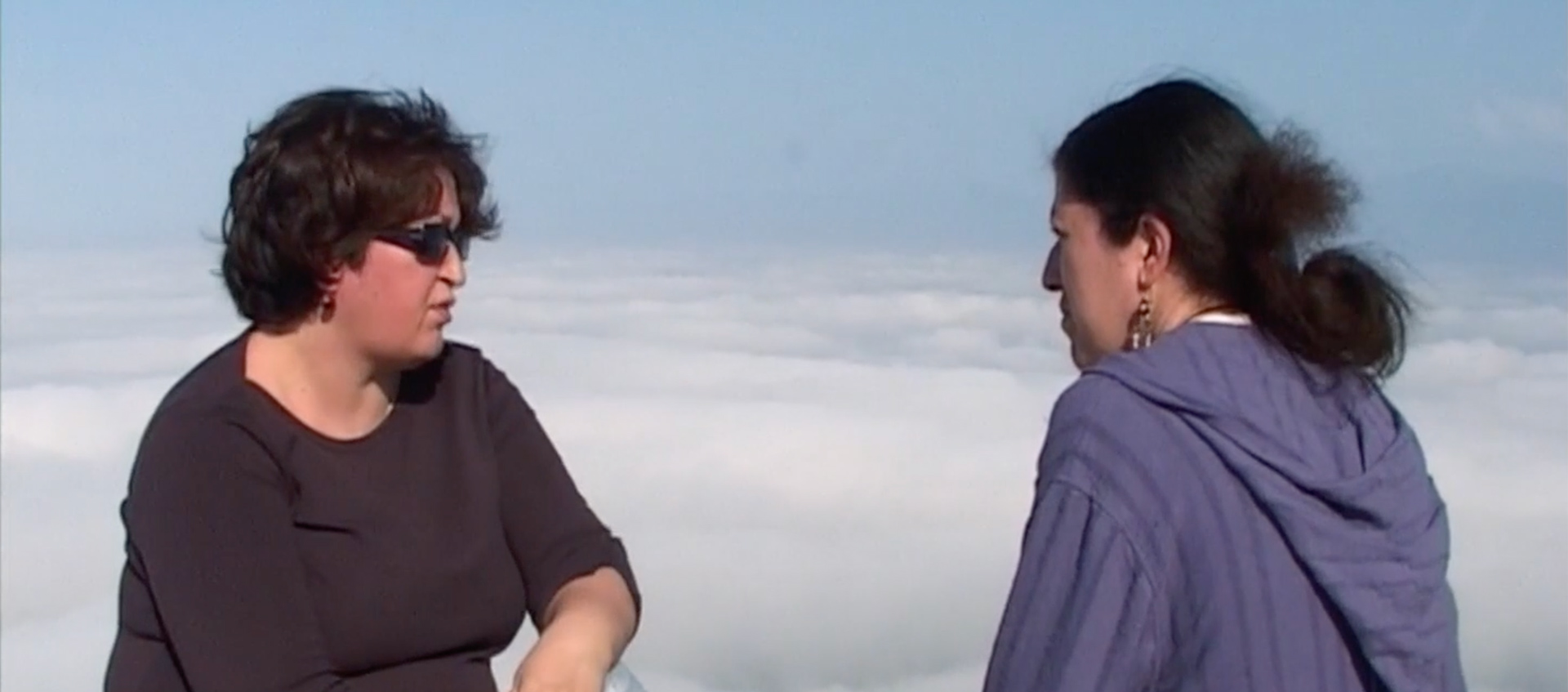 Two women having a conversation on a high altitude with clouds behind in the scene. 
