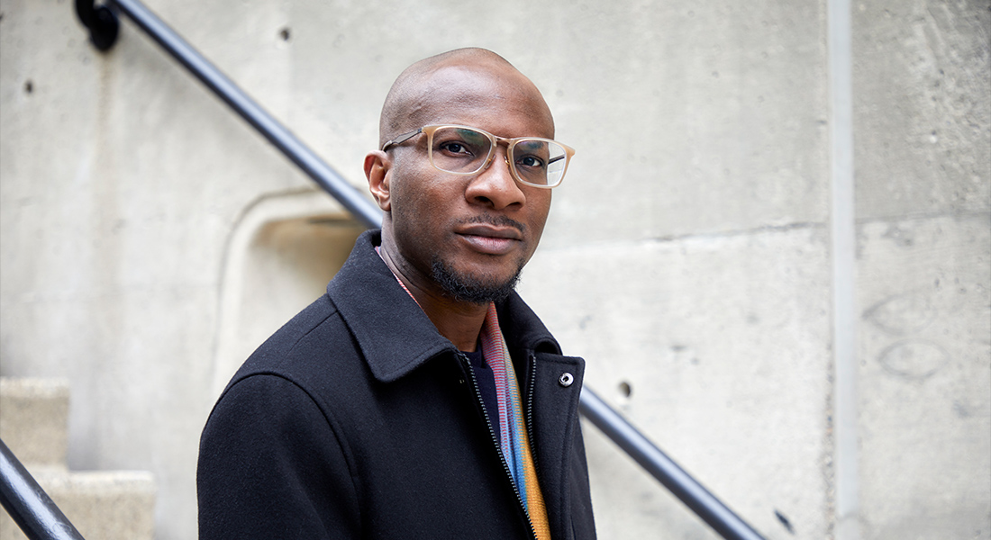 Photo of Teju Cole standing in front of a stairwell, he is wearing eyeglasses and a black jacket