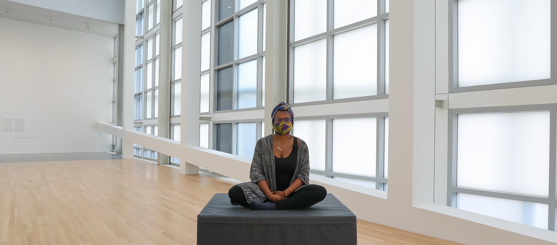 A woman sits on a foam block in the center of an empty gallery space. 