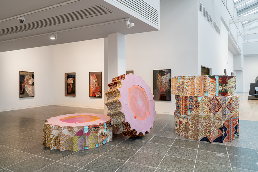 A gallery has framed art on the walls. Large fabric-covered column pieces are stacked on the floor and lean against a column.