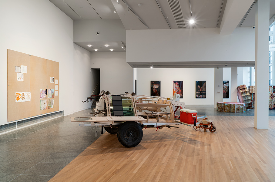 A large artwork made of found objects sits on a gallery floor. Art is on the walls. 