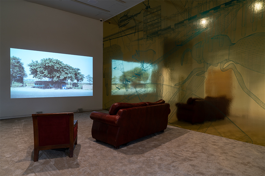 A film is projected onto a wall. A couch and chair sit in front of it. A large vinyl mural is on the wall next to the film.