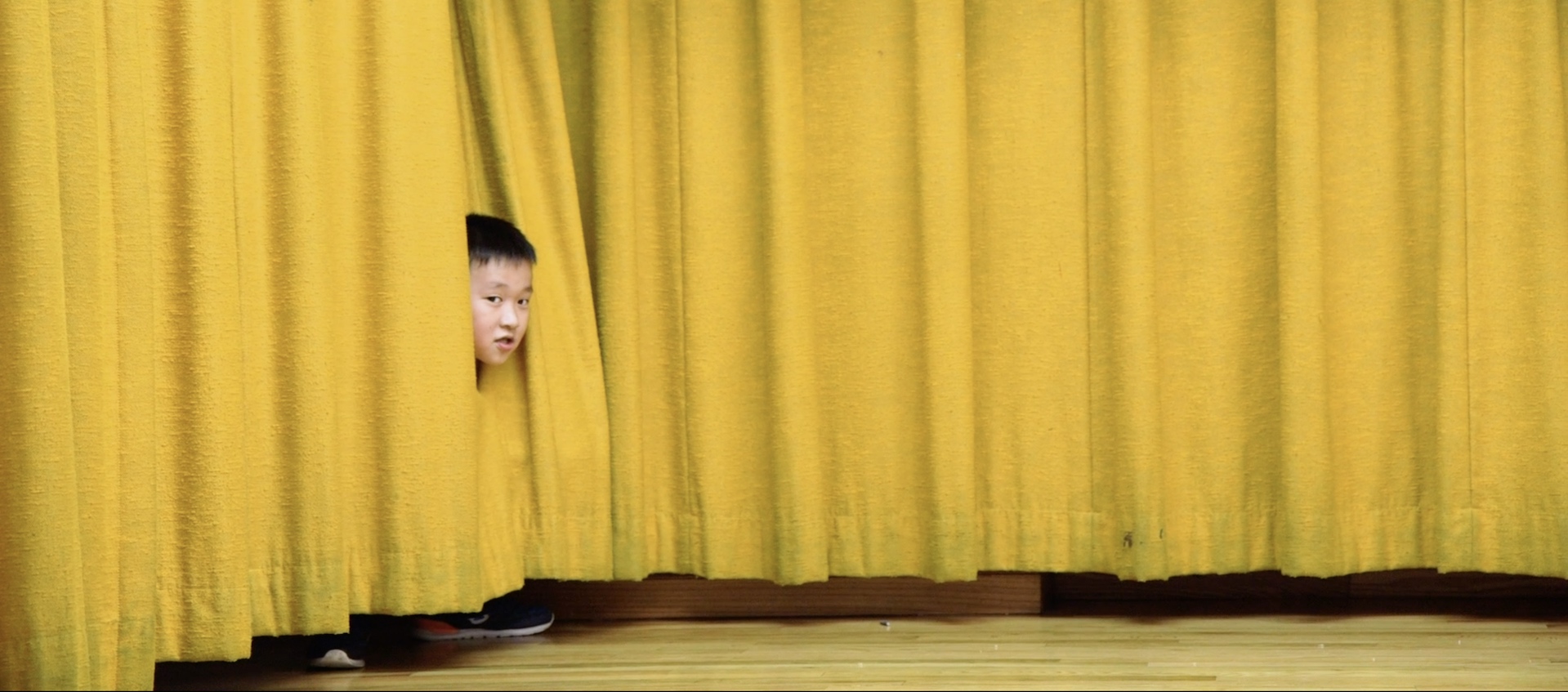 A young boy peeks his head out from behind a big yellow curtain