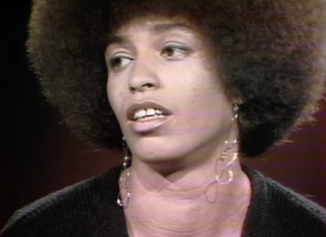 1972 image of Angela Davis making her first national television appearance on the program Black Journal. Davis wears a black shirt and sits in front of a dark red background.