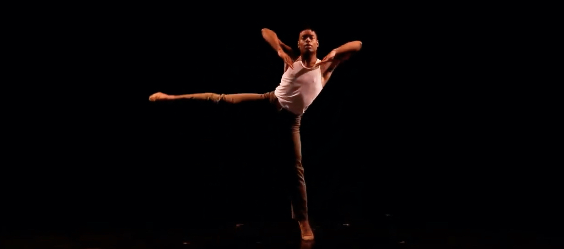 BalletMet Columbus dancer Vincent Harris mid-performance on a darkened stage, with his arms bent and elbows high and his right leg extended straight out to the side