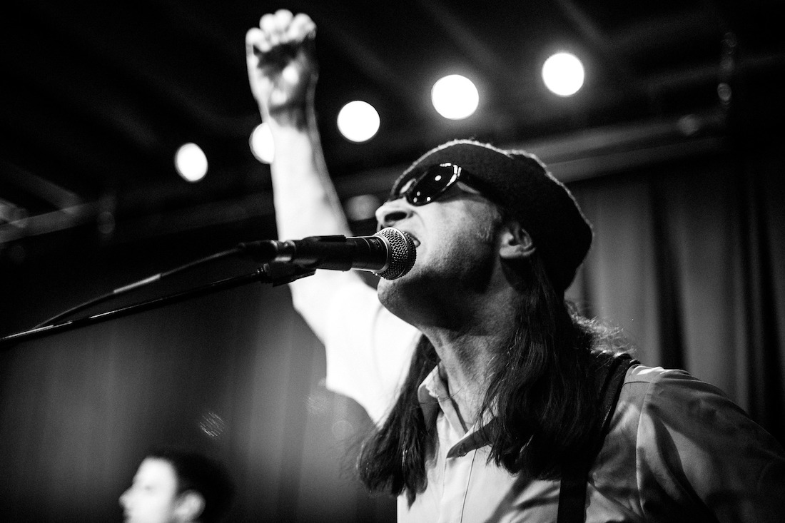 Black and white image of musician Happy Chichester seen in three-quarter profile from the shoulders up, standing on stage wearing a knit hat and sunglasses, his right arm raised with clenched fist; photo by Kris Misevski