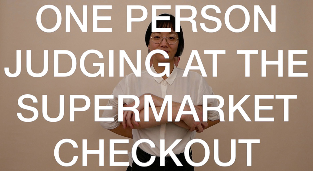Artist Christine Sun Kim looks at the camera against a neutral background with her partner Thomas Mader's arms crossed in front of her (the only part of him visible in the shot). In large white capital letters reads the intertitle (ONE PERSON JUDGING AT THE SUPERMARKET CHECKOUT on top of the pair. 