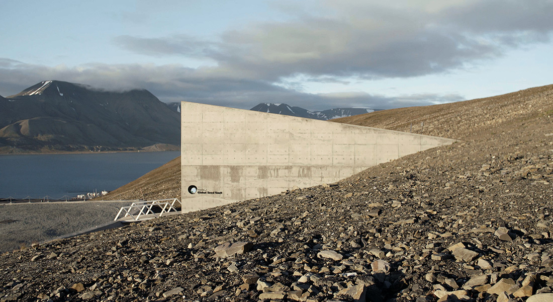 The angled concrete exterior the Global Seed Vault rises out of a rocky shoreline in front of a mountain lake
