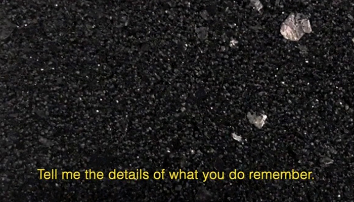 This video still hosts a dark rocky asphalt ground with flecks of mica; it feels like looking down at a road. Open captions in yellow read: “Tell me the details of what you do remember.”