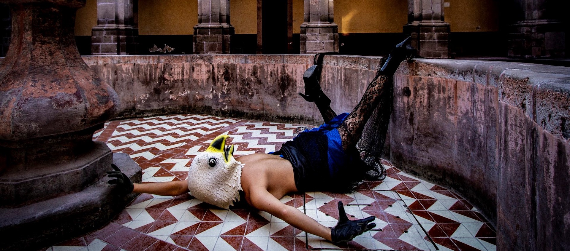 Jessica Bastidas from the group La Pocha Nostra lies face down on a tiled floor in the middle of a round space filled with aged columns, topless with a lacy skirt, tights and high heels, a large eagle mask covering her head