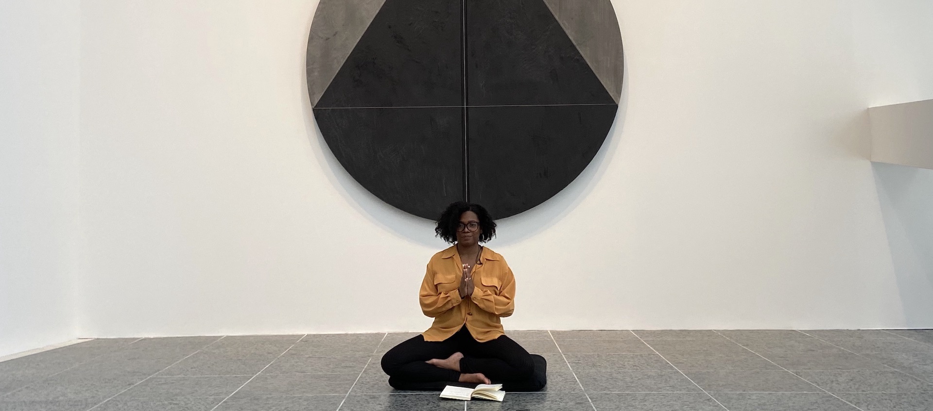 Monique McCrystal sits on the tile floor of the Wexner Center for the Arts galleries with her hands in prayer pose, underneath a round abstract painting in black and gray by Torkwase Dyson