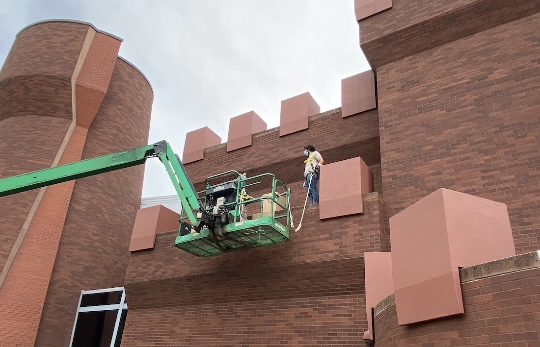 Wexner Center for the Arts preparators Dave Dickas and James-David Mericle stand on a mechanical lift, installing crenellations for Chris Burden's Wexner Castle onto the exterior of the center building