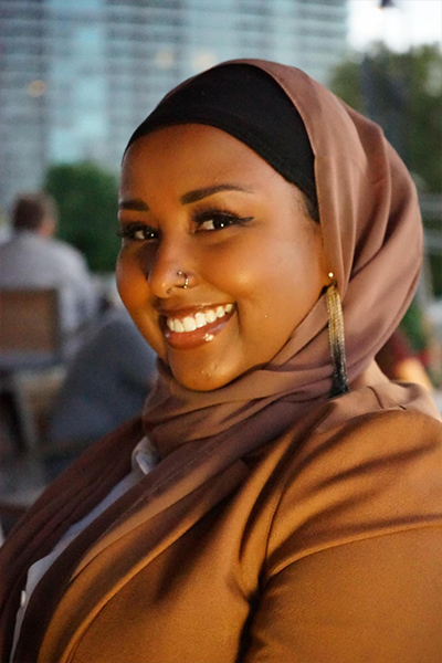 A color photo of Ruun Nuur in a brown top and head scarf. Nuur is smiling at the camera.