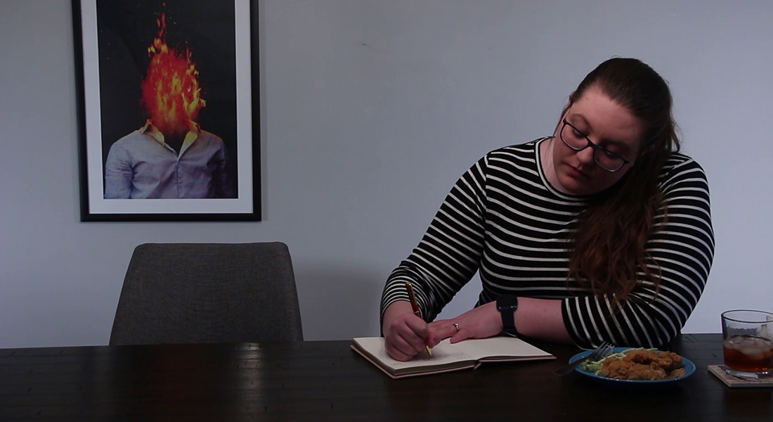 A color film still of a person in glasses and a striped shirt writing in a journal; a plate of food and a glass is at their right; behind the person is a picture of another person with their head engulfed in flames