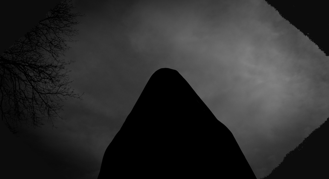 A shadowy image looking up at a cloudy night sky with bare tree branches visible to the left and the outline of a rectangular structure seen from an angle in the center