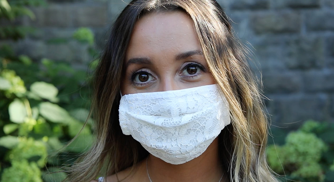 A color still of a person in a lace face mask with their eyes visible above; a stone wall and low plants are in the background