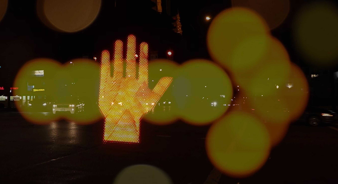 A color film still superimposing an illuminated hand symbol from a traffic sign over the lit arrow from another traffic sign, shot out of focus, all against cars driving along a city street at night
