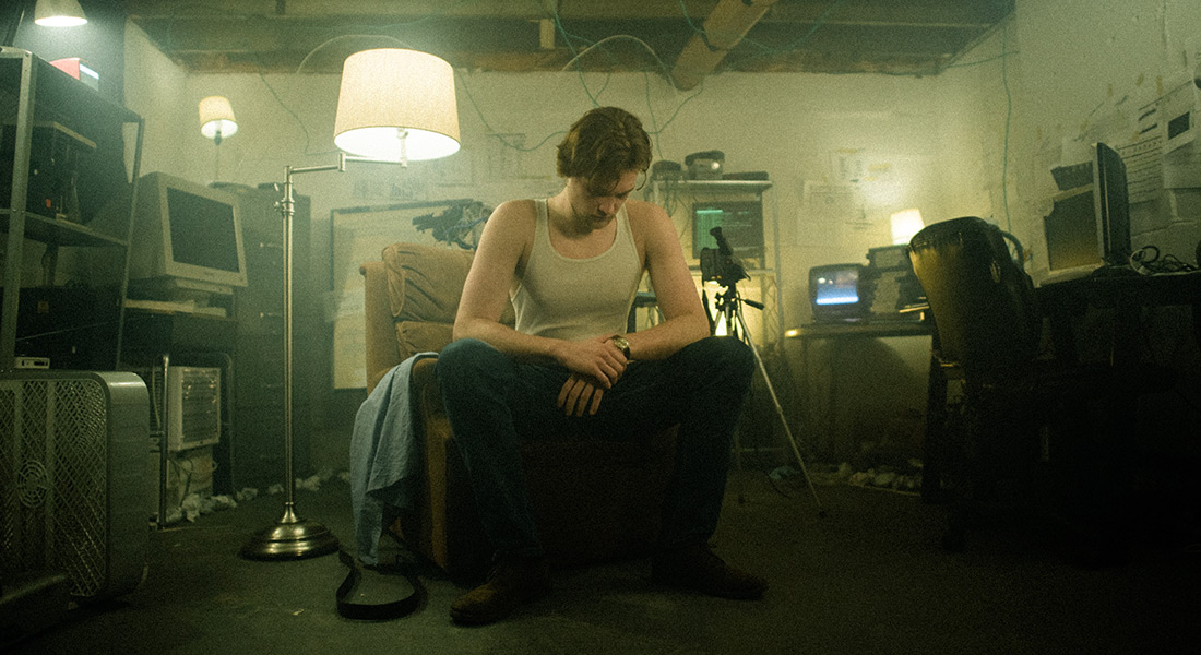 A color film still showing a person in a white tank top seated, head down, on a chair in a cluttered basement filled with lamps, computer monitors, and furniture