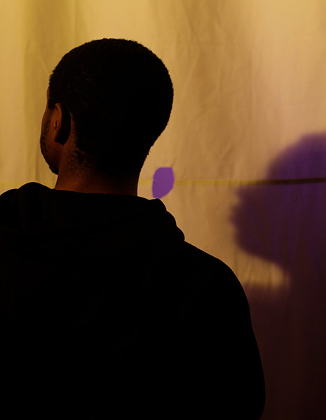 A film still in color showing a person seen from the back in silhouette; the person stands against a sheet with a purple spot in the middle