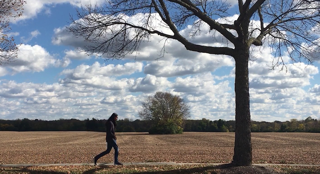 A long-haired young person in a purple shirt and blue jeans walks along on a path in front of a deserted plowed field under a partly cloudy sky