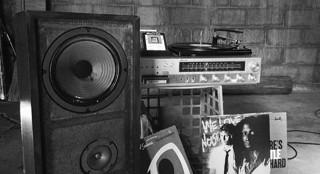 A black-and-white still showing a close-up of a late-1970s era home stereo system sat atop a milk crate with a few vinyl records stood in front of the grate; to the left of the turntable is a large speaker with no grille; in the background is an unpainted cinder-block basement wall