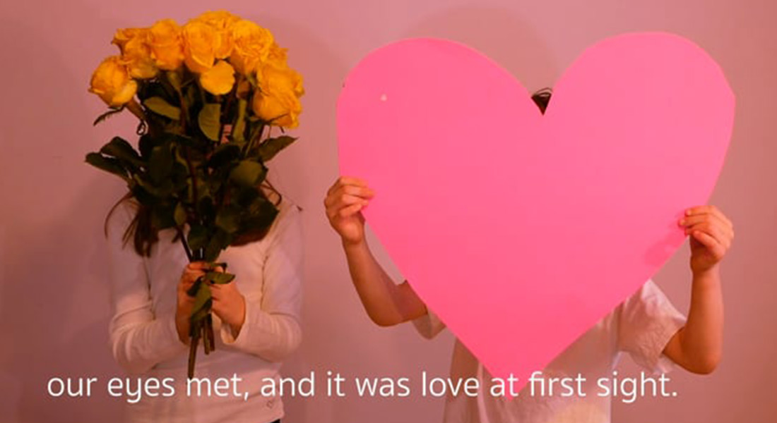 A color still showing two people against a pink background; the person on the left hides their face with a bouquet of yellow flowers, and the person on the right holds up a large flat pink heart in front of theirs