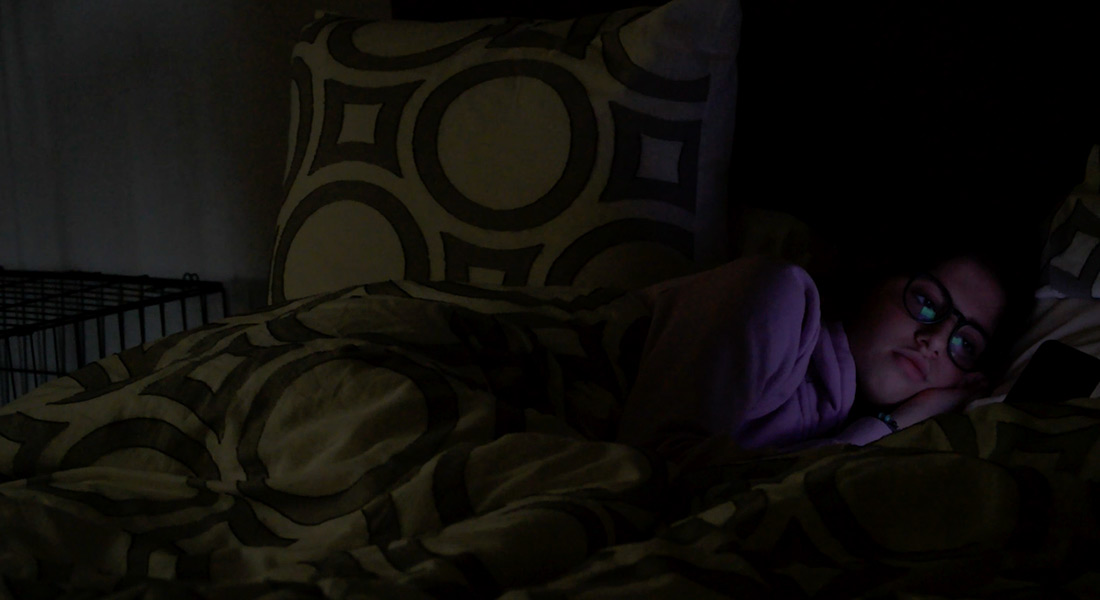A color still showing a person in bed under a patterned yellow duvet in a very dimly lit room; the light of a screen reflects in the person's glasses
