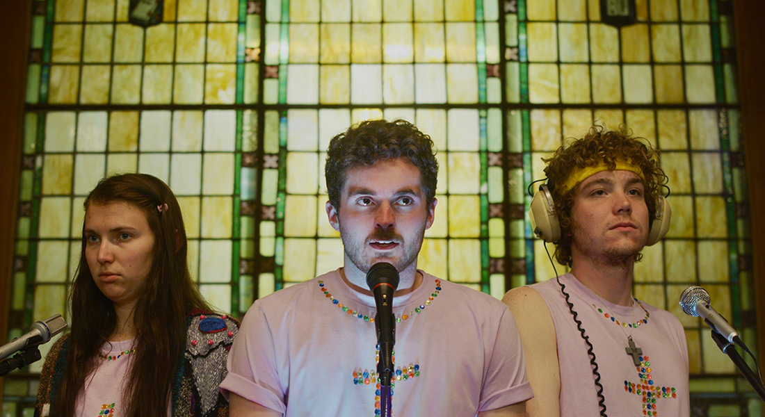 A color still of three young people standing in front of microphones in front of a stained-glass window wearing white shirts with jeweled crosses and necklines