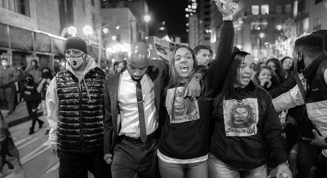 A black-and-white still showing people marching through the streets of Columbus at night; two people in the foreground wear shirts with the same drawn portrait of Casey Goodson Jr. and the words "Justice for Casey"