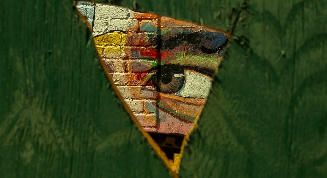A color close-up of a mural of a person's face painted on a brick wall; the eye is framed by a triangular hole roughly cut into a piece of wood painted green