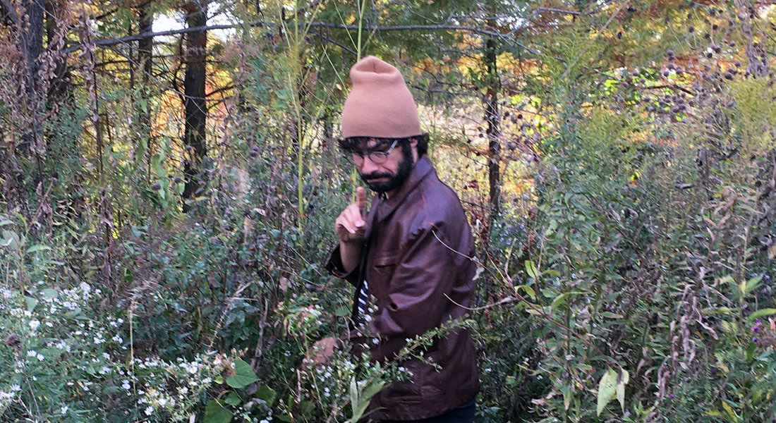 A bearded person in glasses, a beige hat, and leather jacket gestures amid a forest