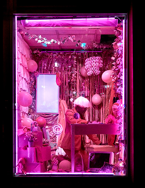 A vertical image of a room in a pink tint. Raja Feather Kelly is seated and the wall behind them is covered in tinsel and balloons.