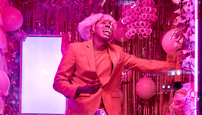 A hot pink image with Raja Feather Kelly in the center of the frame standing on a stage. Kelly is in a pink jacket and wig with arm outstretched. Kelly appears to be singing and behind Kelly are balloons and tinsel covered backdrop. 