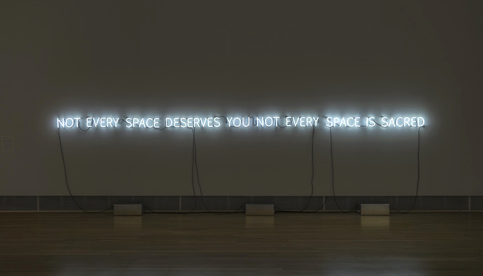 "Not every space deserves you not every space is sacred" in white neon lettering on a white wall in a darkened gallery
