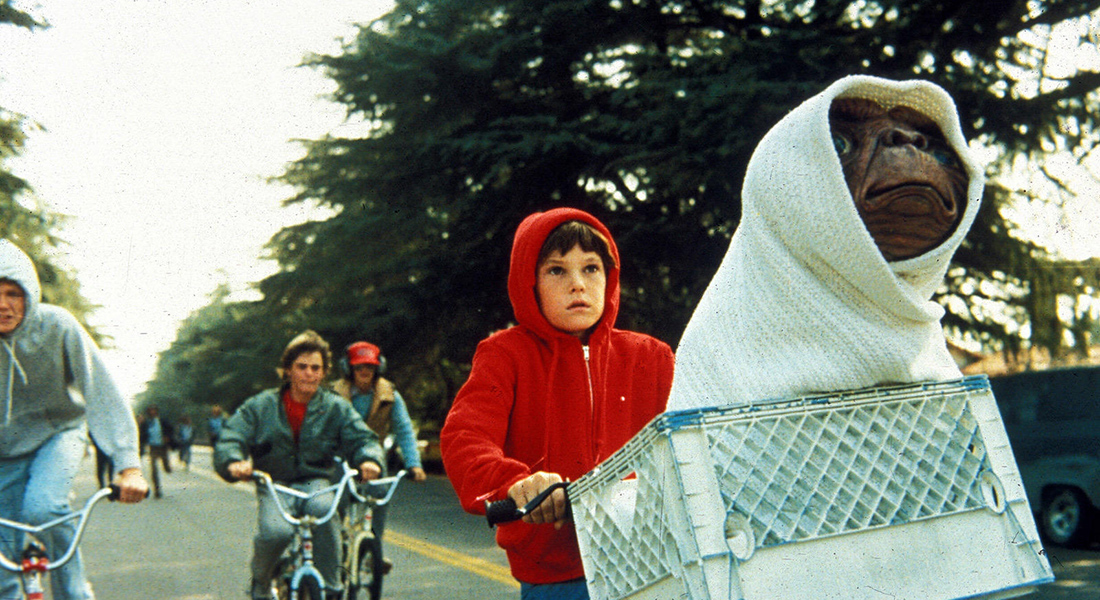 A boy in a red hoodie (Henry Thomas) rides a bike with an alien in a milkcrate on his handlebars with several other riders behind them